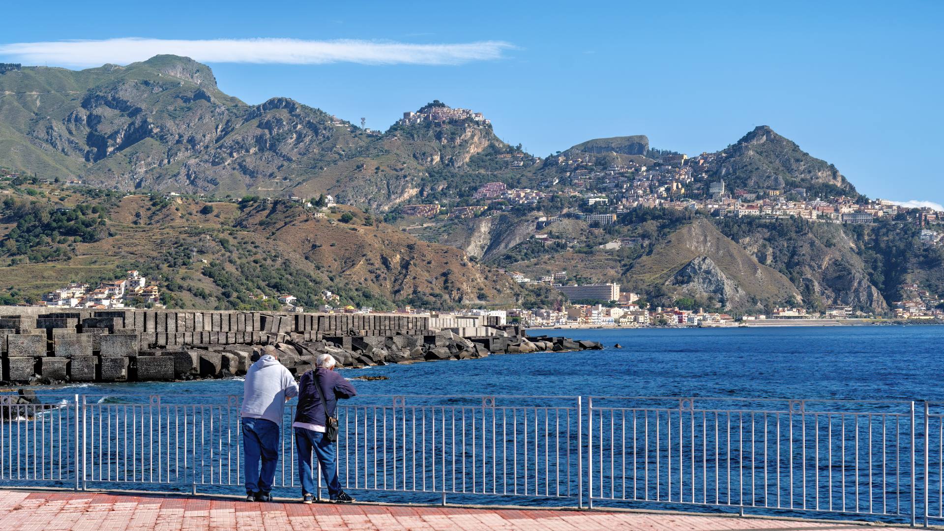 Blick von Giardini-Naxos nach Taormina: Am Ufer sieht man die Ausläufer von Giardini-Naxos, auf halber Höhe liegt Taormina und ganz oben thront der kleine Ort Castelmola